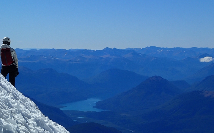On the left side of the photo, a person wearing safety gear stands on a snowy incline, looking out over a vast mountainous landscape and a blue body of water. The mountains appear blue under the blue sky. 